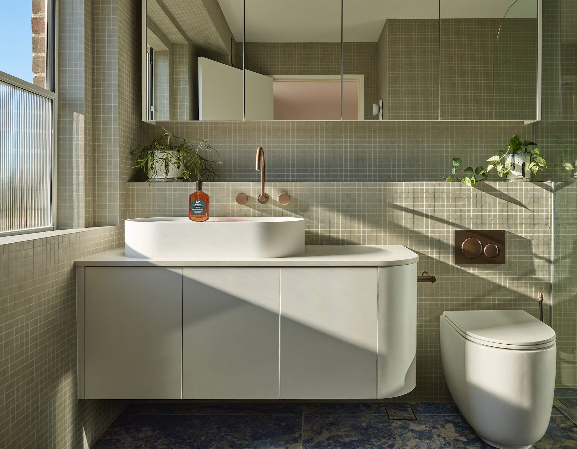 Bathroom with sage-green mosaic tiles, a curved floating vanity with a white basin and bronze-toned tapware in afternoon light