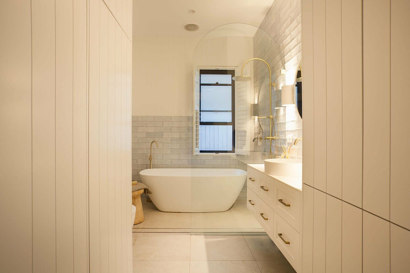 Bathroom with a freestanding bath, white vertical panelling, an arched glass screen and brass tapware