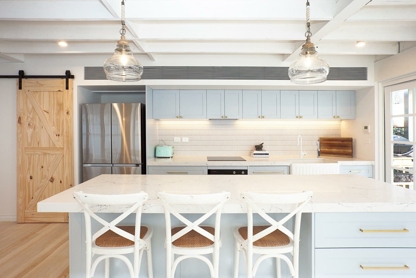 Kitchen with pale blue cabinetry, a white island bench with three stools and glass pendant lights