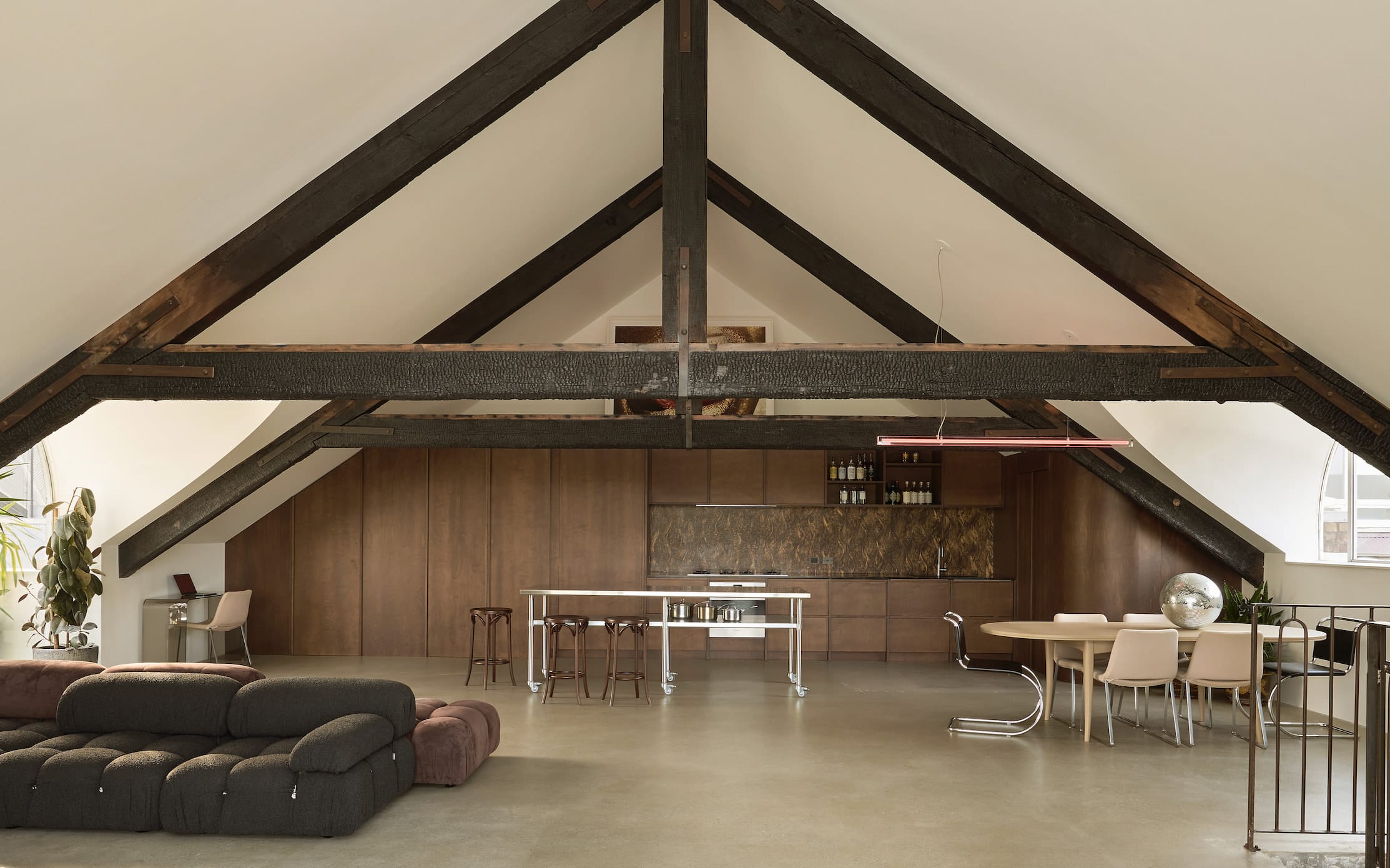 Open-plan loft living space beneath dark exposed roof trusses, with timber-panelled joinery along the back wall, a steel kitchen island and a dining table