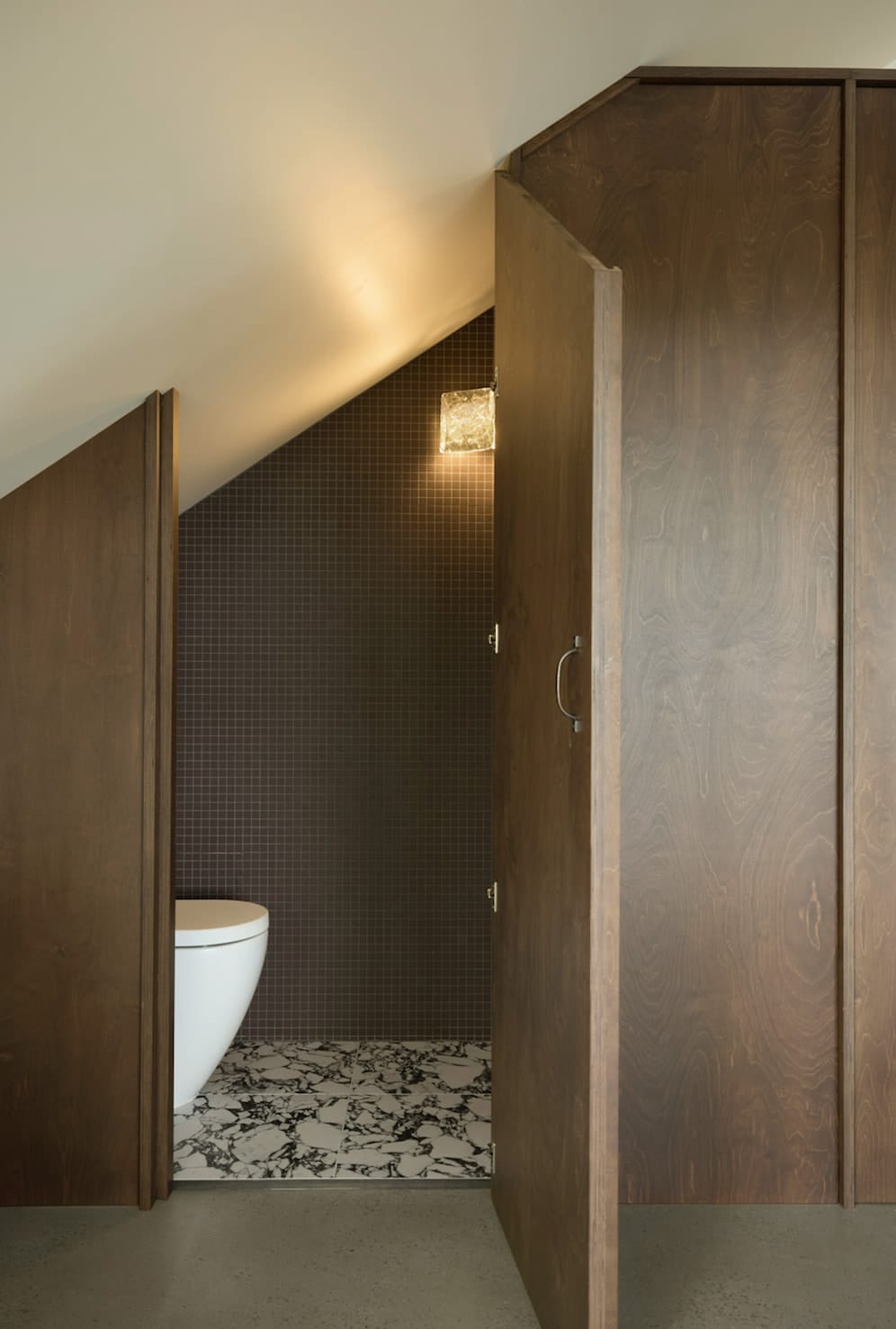 Powder room tucked under the sloping roof behind a timber door, with dark mosaic wall tiles and a black-and-white patterned floor