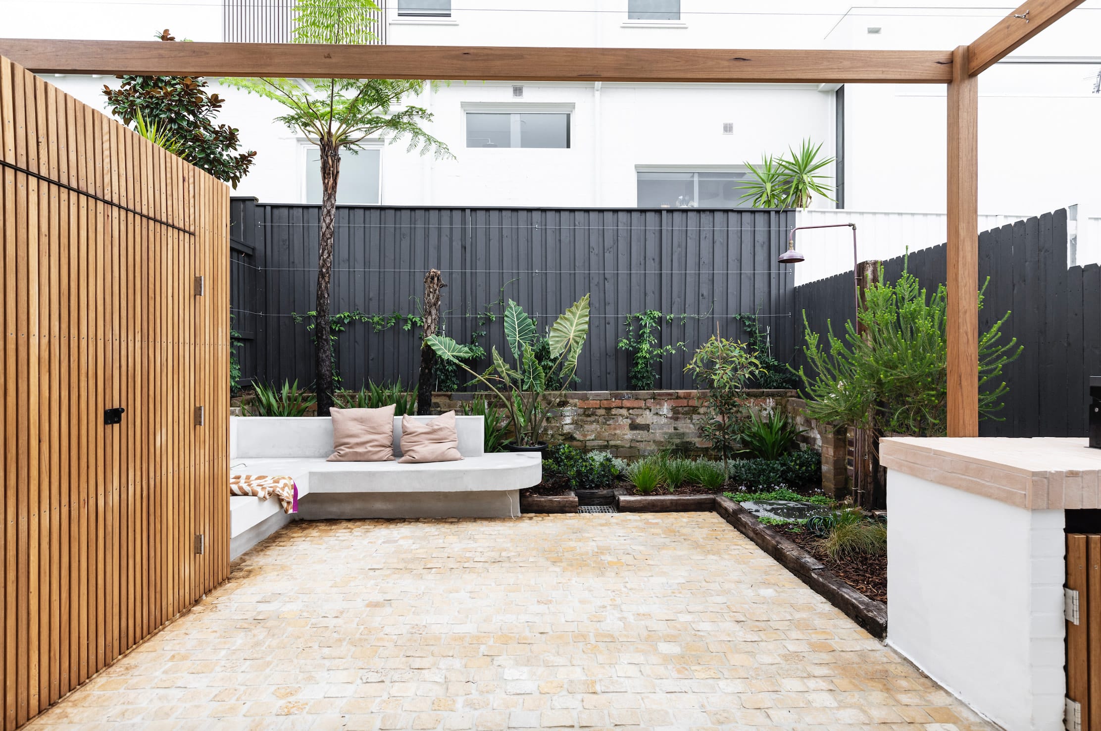 Rear courtyard with stone cobble paving, a built-in rendered bench, a timber pergola beam and charcoal-painted fencing