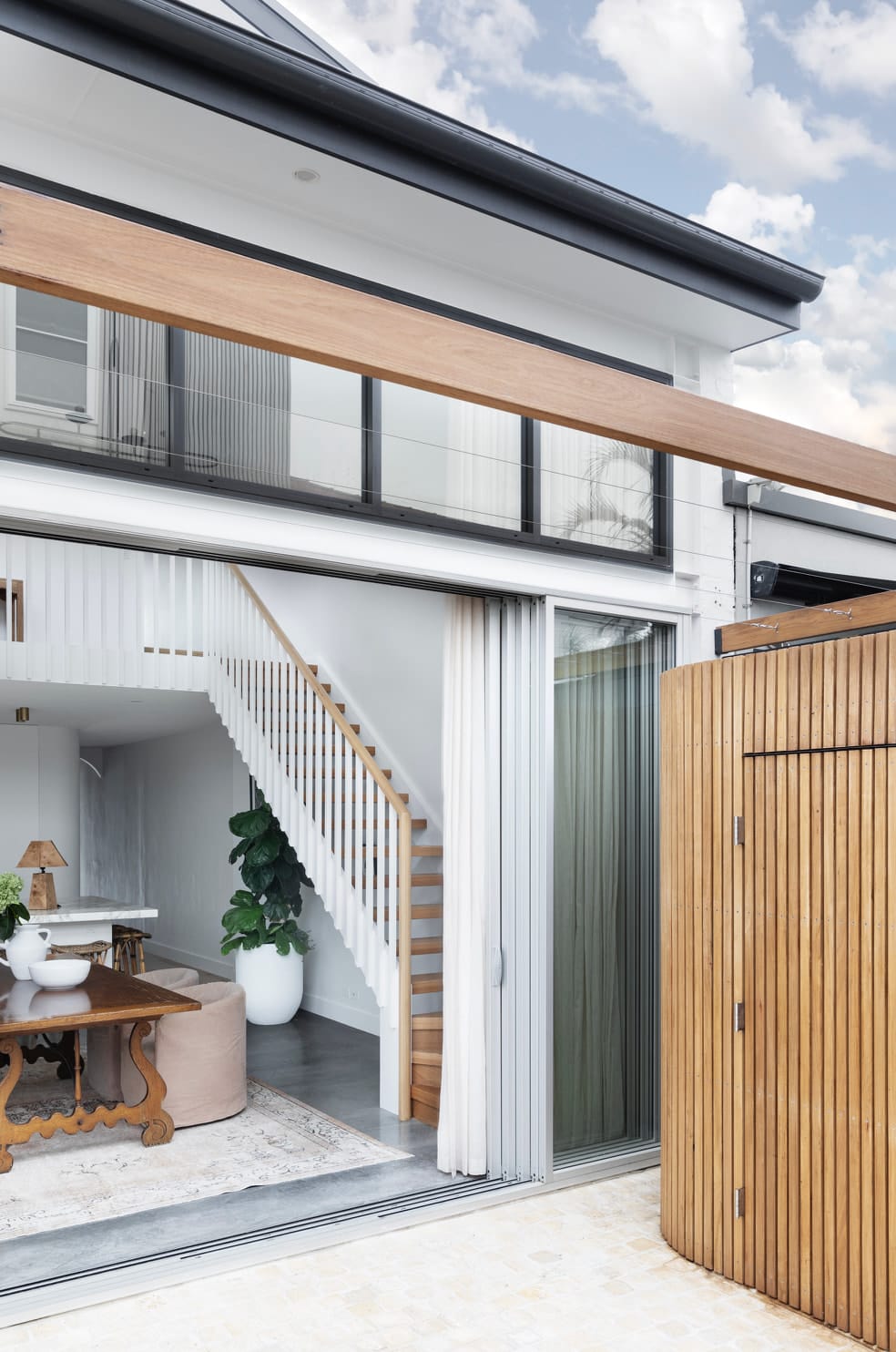 Back of the house with wide glass doors opening to a living area and timber stair, beside a curved timber-batten enclosure